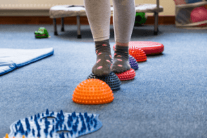 A child wearing gray leggings and polka dot socks balances on colorful textured stepping stones in a playroom with a blue carpet, reflecting the engaging environments often used in autism therapy near Chester County PA.