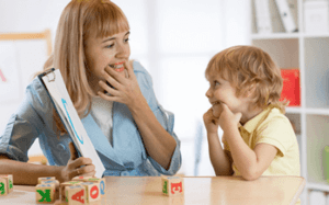 A woman and a young child sit at a table with colorful letter blocks, both touching their faces and smiling at each other, possibly practicing speech or communication exercises as part of autism services near Montgomery County PA.