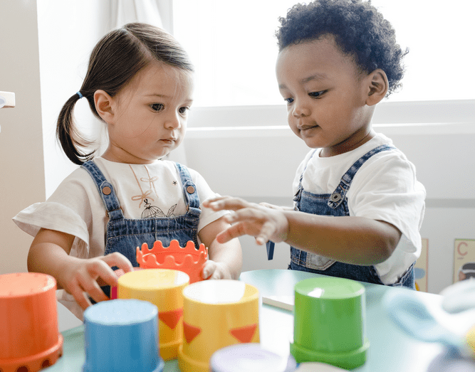 Two young children wearing overalls play together at a table, stacking colorful toy cups and examining an orange block. This joyful moment highlights the benefits of autism therapy near Chester County PA. Natural light fills the room through a window.
