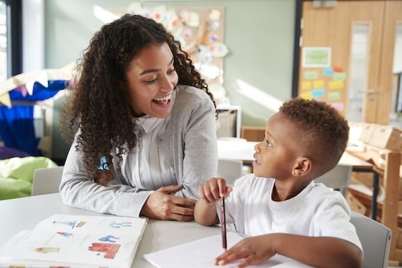 A woman and a young boy sit at a table in a classroom, smiling at each other. The boy holds a pencil and notebook, and there are drawings on the table—capturing a joyful moment during autism therapy near Chester County PA.