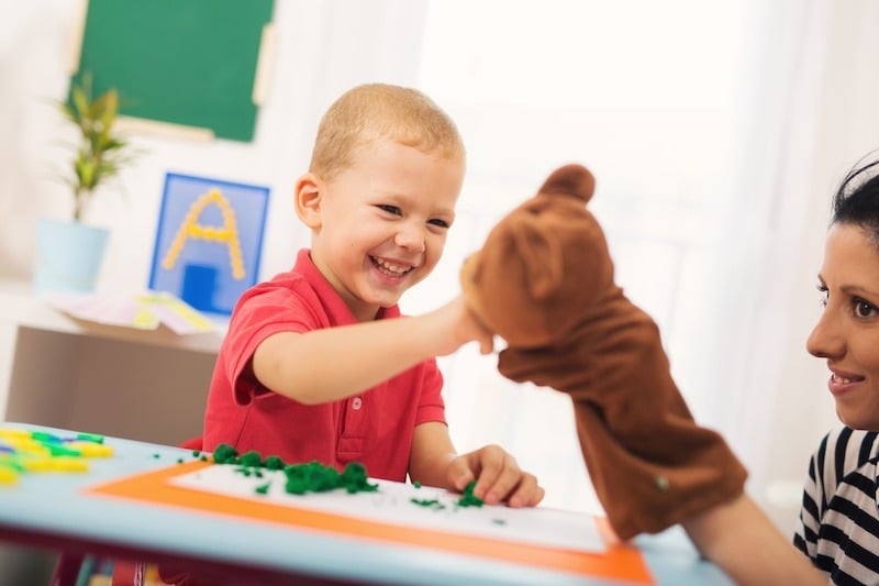 A young boy smiles and plays with a brown teddy bear puppet held by an adult at a table covered with green playdough and colorful toys in a bright room offering ABA therapy near Philadelphia PA.