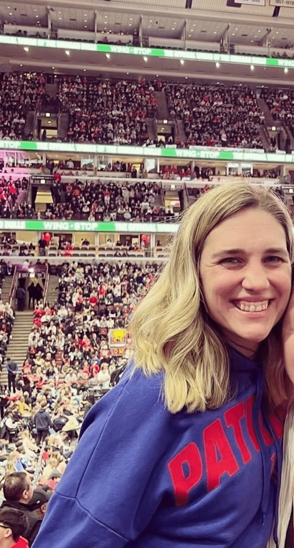 A smiling woman in a blue sweatshirt with red lettering stands in the foreground at a crowded sports arena; rows of fans fill the seats behind her, highlighting the vibrant community near ABA therapy near Philadelphia PA.