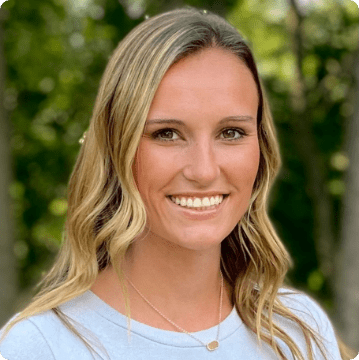 A young woman with long, wavy blonde hair smiles at the camera. She is wearing a light blue top and a necklace, standing outdoors with green trees blurred in the background—an inviting image for those seeking autism therapy near Chester County PA.