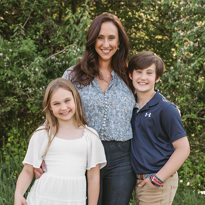 A woman stands outdoors with two smiling children, a girl in a white dress and a boy in a navy polo shirt, enjoying the fresh air—perfect for families seeking autism services near Montgomery County PA.