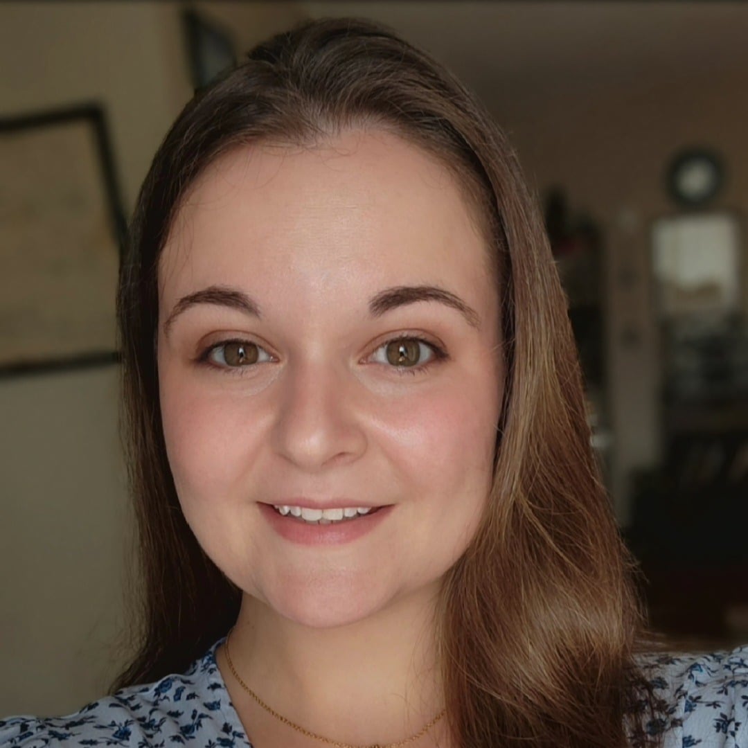 A young woman with long brown hair and brown eyes smiles at the camera. She is wearing a blue patterned top and a delicate necklace, with a softly blurred indoor background—perfect for promoting autism therapy near Chester County PA.
