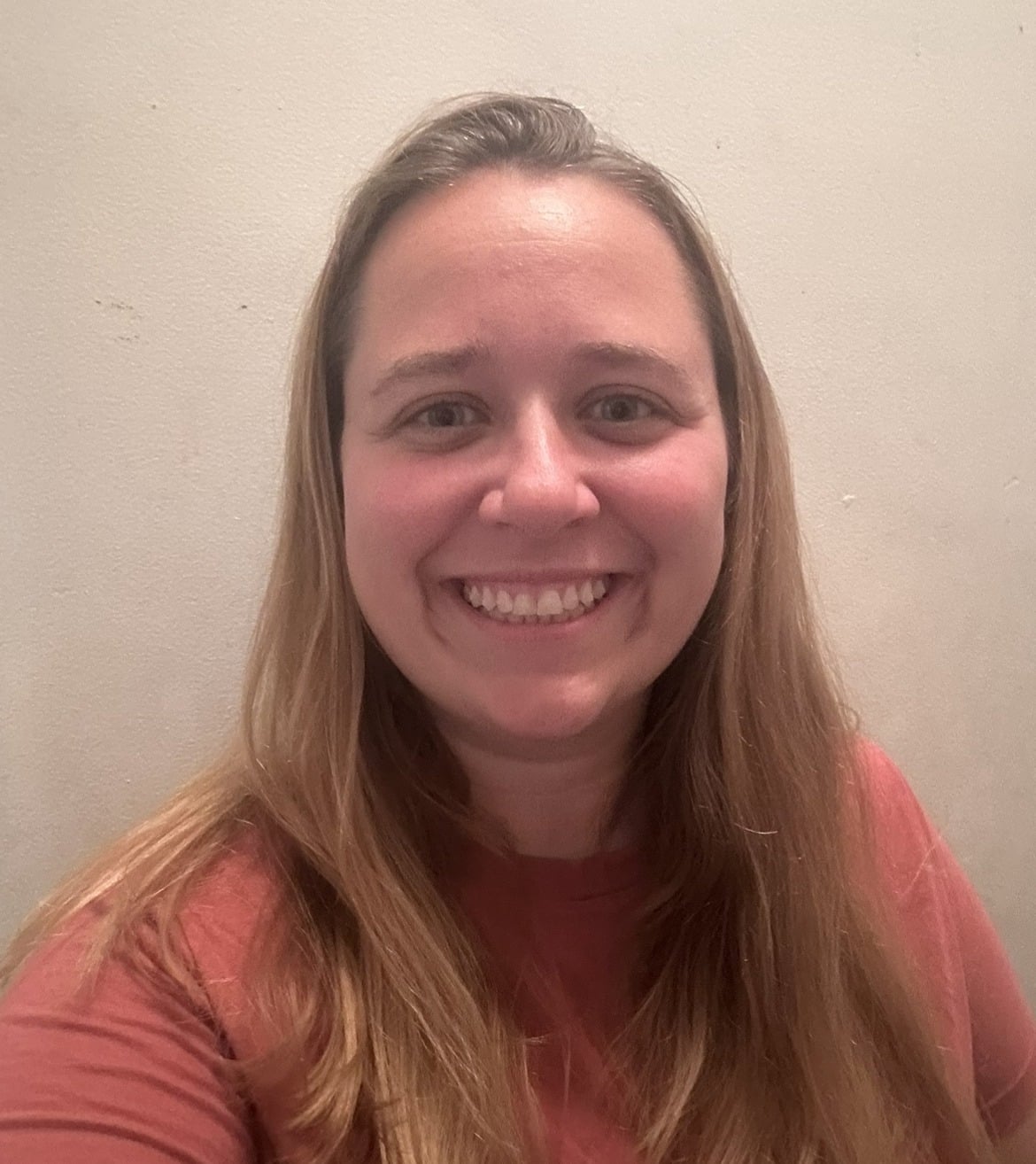 A person with long, light brown hair smiles at the camera. Wearing a reddish-orange shirt, they stand before a plain, light-colored wall—representing hope inspired by autism therapy near Chester County PA.
