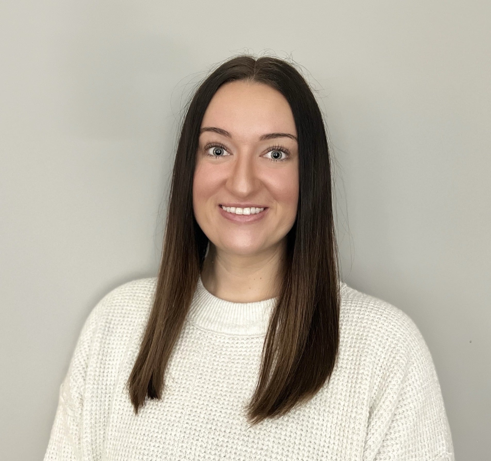 A woman with straight brown hair, wearing a white knit sweater, smiles at the camera against a plain light gray background, representing autism therapy near Chester County PA.