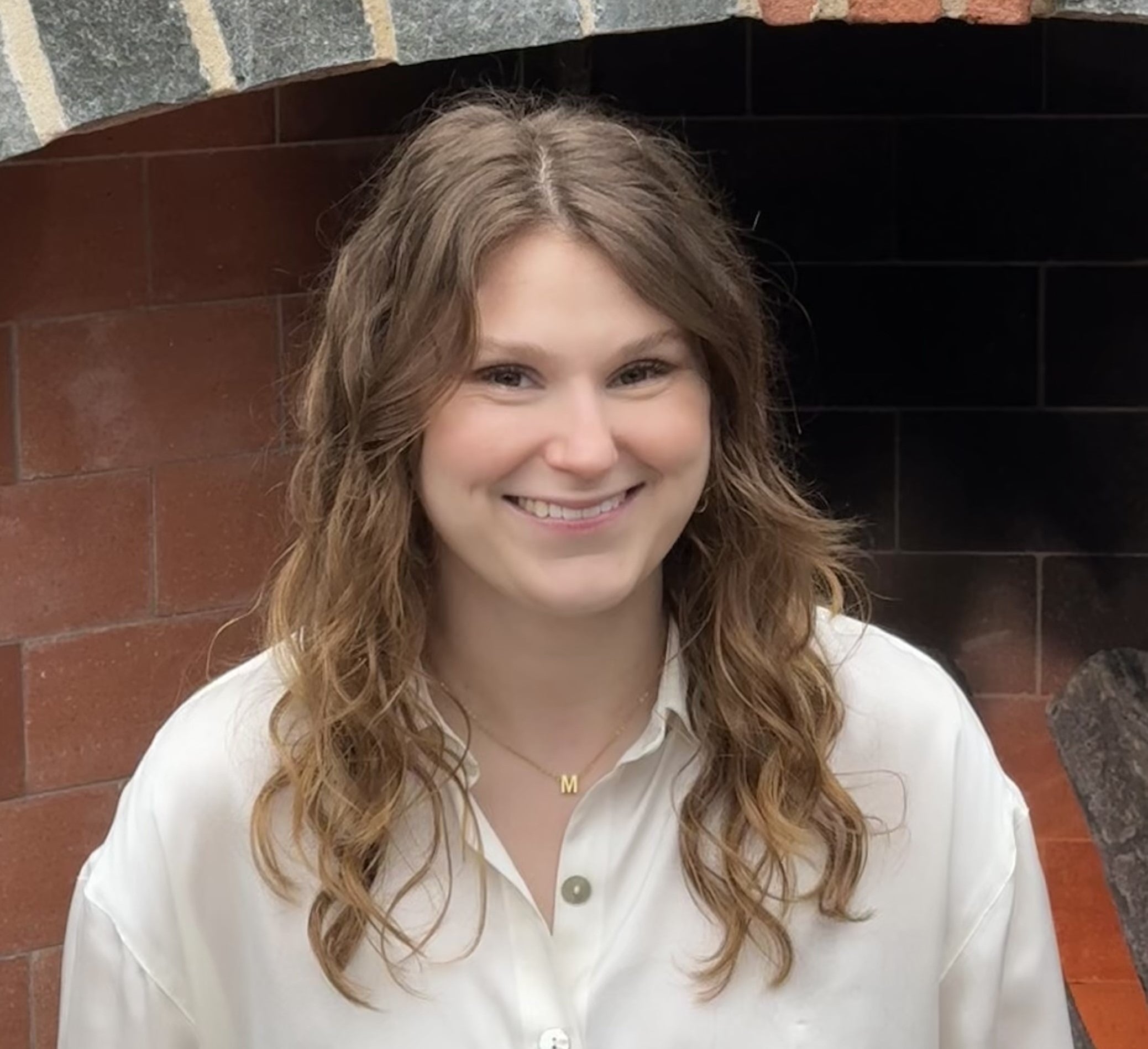 A woman with wavy brown hair, wearing a white button-up shirt and a gold "M" necklace, smiles in front of a brick and stone fireplace—an inviting face for those seeking ABA therapy near Philadelphia PA.