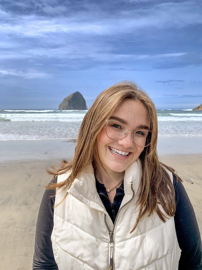 A smiling woman with glasses and a white vest stands on a sandy beach with waves and a large rock formation in the background under a blue sky, reflecting the calm support of autism therapy near Chester County PA.