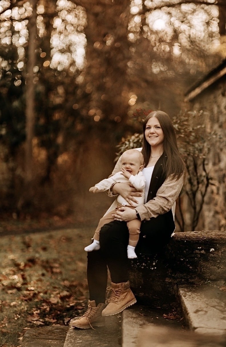 A woman sits on outdoor stone steps holding a smiling baby on her lap. Both are dressed in casual fall clothes, surrounded by autumn trees and soft, warm sunlight—perfect for families seeking autism therapy near Chester County PA.