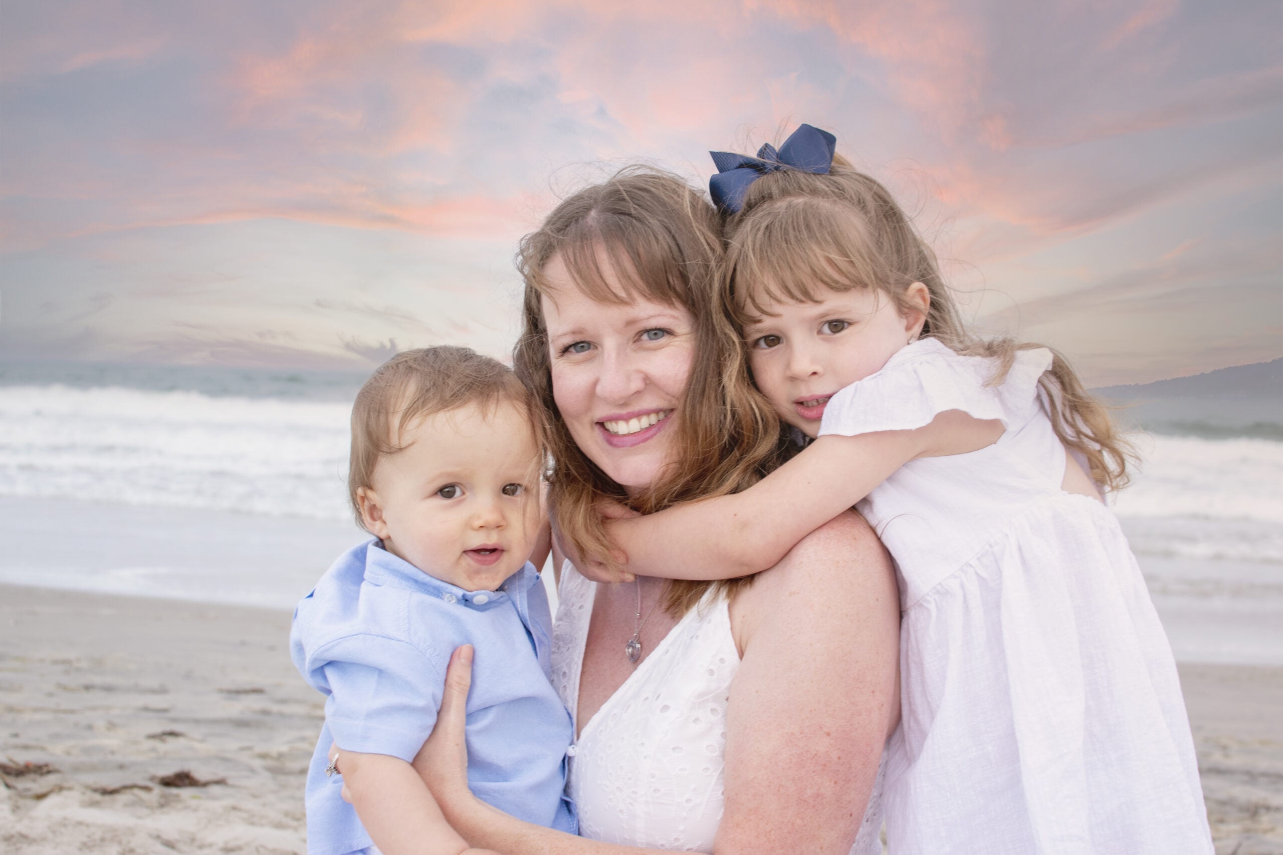 A woman smiles while hugging two young children on a sandy beach at sunset, capturing a joyful moment that families seeking autism therapy near Chester County PA cherish. Waves and a colorful sky complete the heartwarming scene.