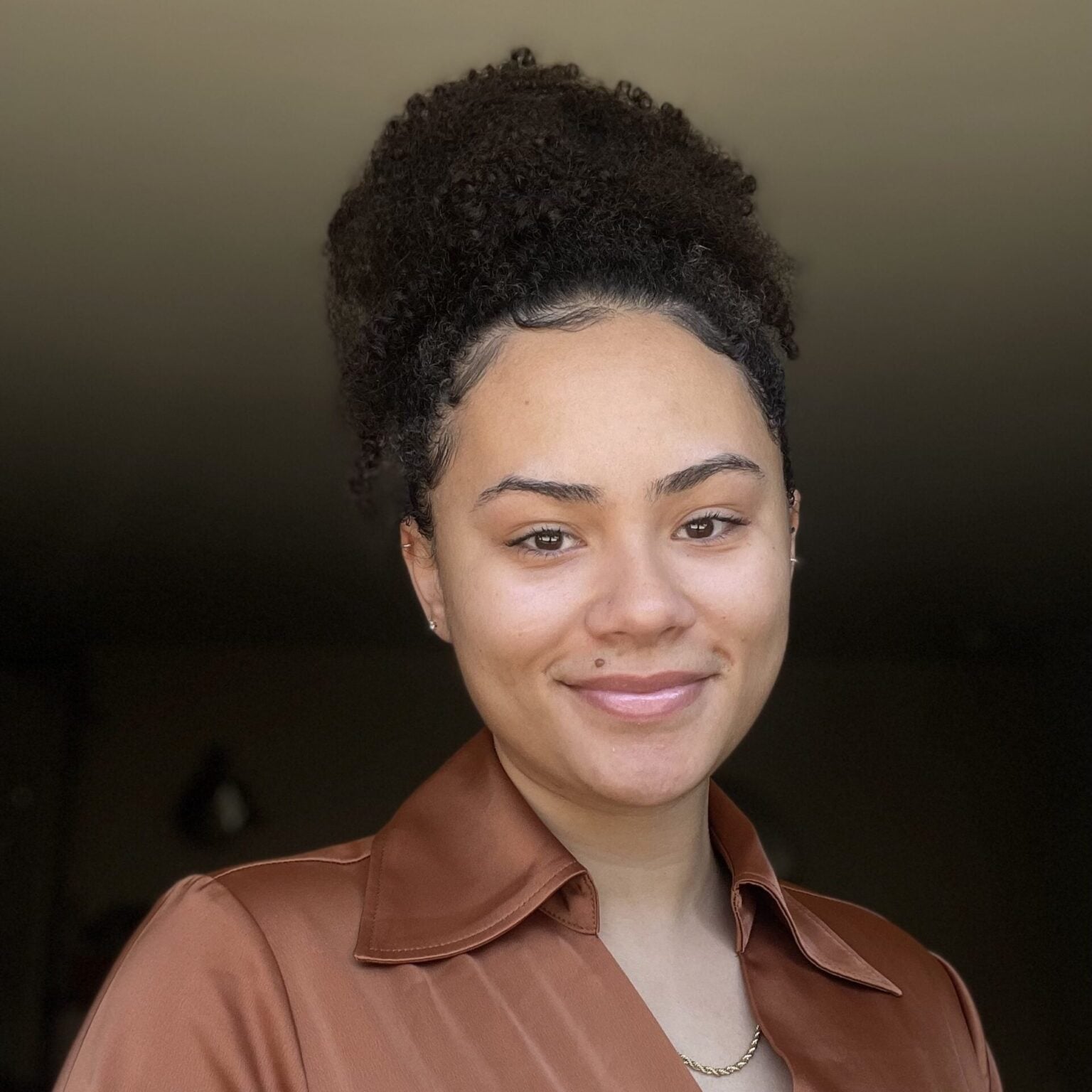 A young woman with curly hair pulled into a high puff smiles softly. She wears a brown satin shirt, gold necklace, and has a small mole above her lip. The softly lit background brings warmth, echoing the care of autism therapy near Chester County PA.
