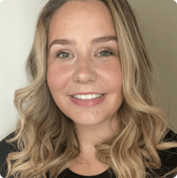 A woman with long, wavy blonde hair smiles at the camera. Wearing a black top and natural makeup, she stands against a plain light background, representing autism services near Montgomery County PA with warmth and approachability.