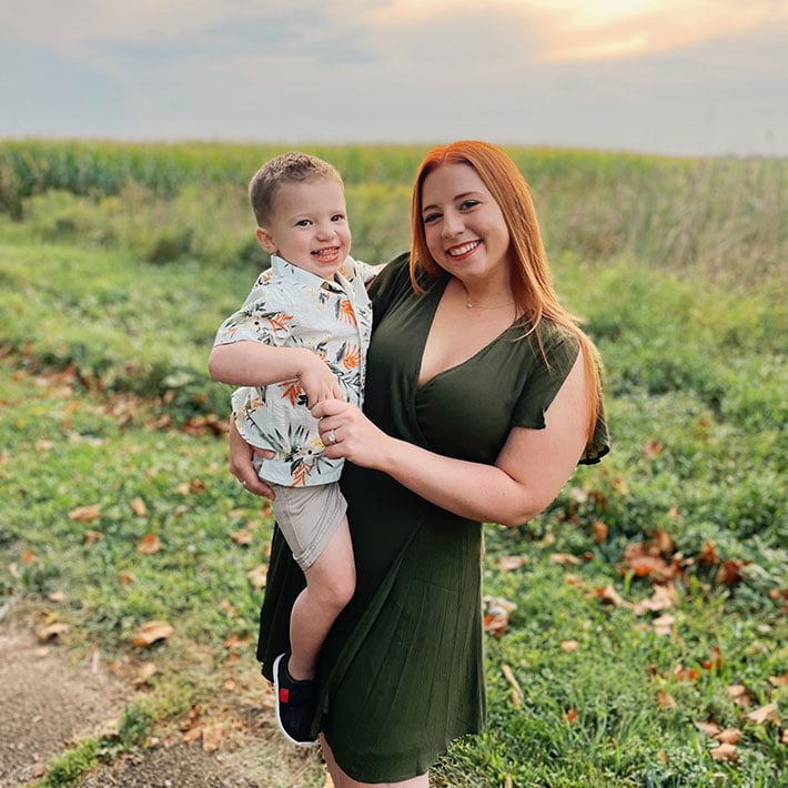 A woman in a dark green dress smiles while holding a young boy wearing a floral shirt and shorts. They stand outside on a grassy path with green fields and a sunset sky, enjoying time together after autism therapy near Chester County PA.