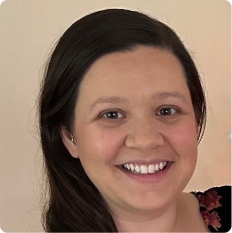 A woman with long dark hair and a friendly smile looks at the camera. She is wearing a dark top with a floral pattern, set against a plain beige background—representing compassionate autism therapy near Chester County PA.