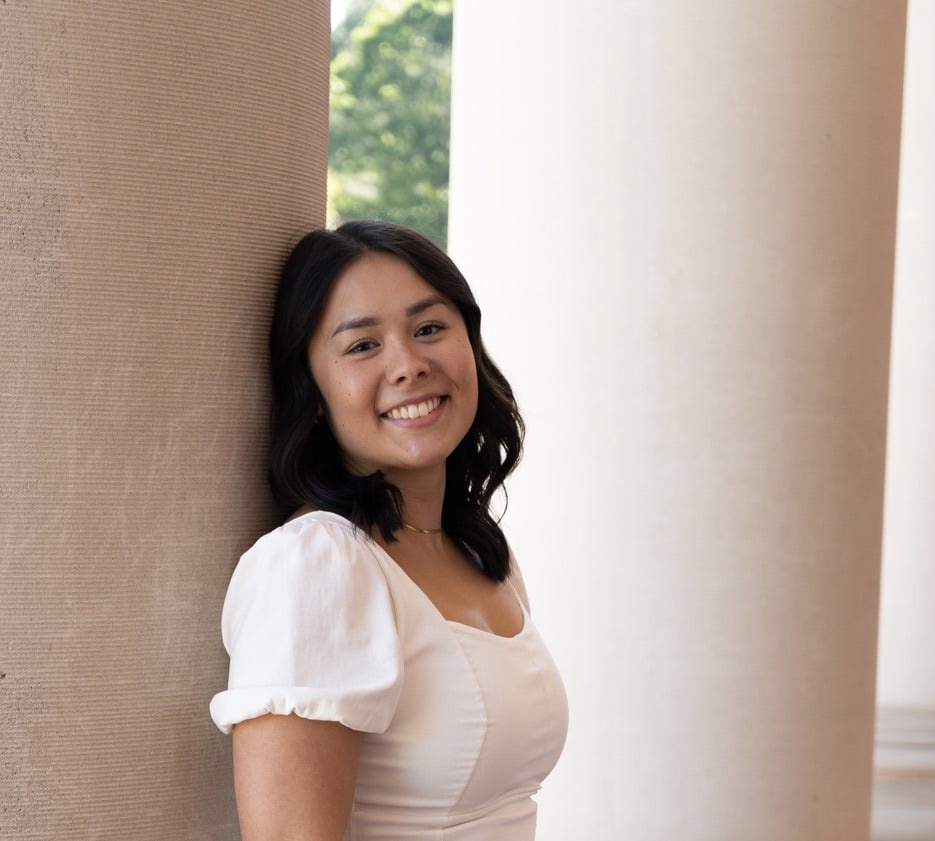 A young woman with medium-length dark hair, wearing a white dress, smiles while leaning against a large column. The background includes another column and greenery near Chester County PA, known for quality ABA therapy.