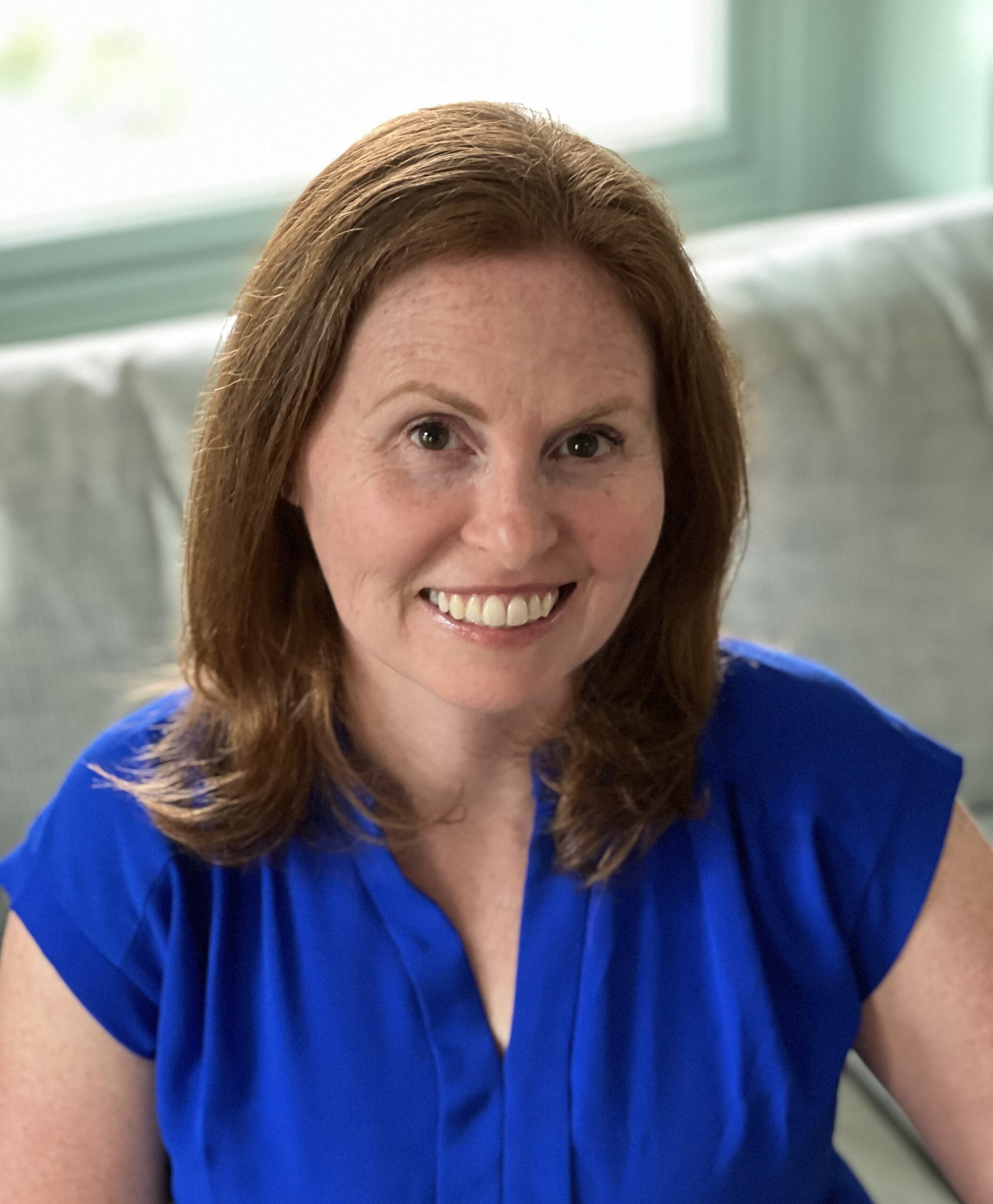 A woman with shoulder-length reddish-brown hair, wearing a bright blue blouse, smiles while sitting on a light gray sofa with a window in the background, representing quality ABA therapy near Philadelphia PA.
