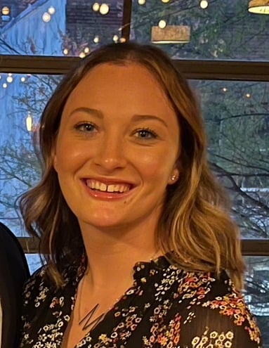 A woman with shoulder-length blonde hair smiles at the camera. She is wearing a black floral top and a necklace, standing indoors with a window and string lights in the background.
