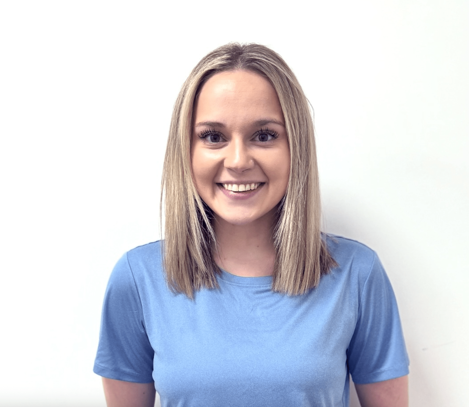 A young woman with straight, shoulder-length blonde hair is smiling at the camera. She is wearing a light blue t-shirt and is standing against a plain white background.