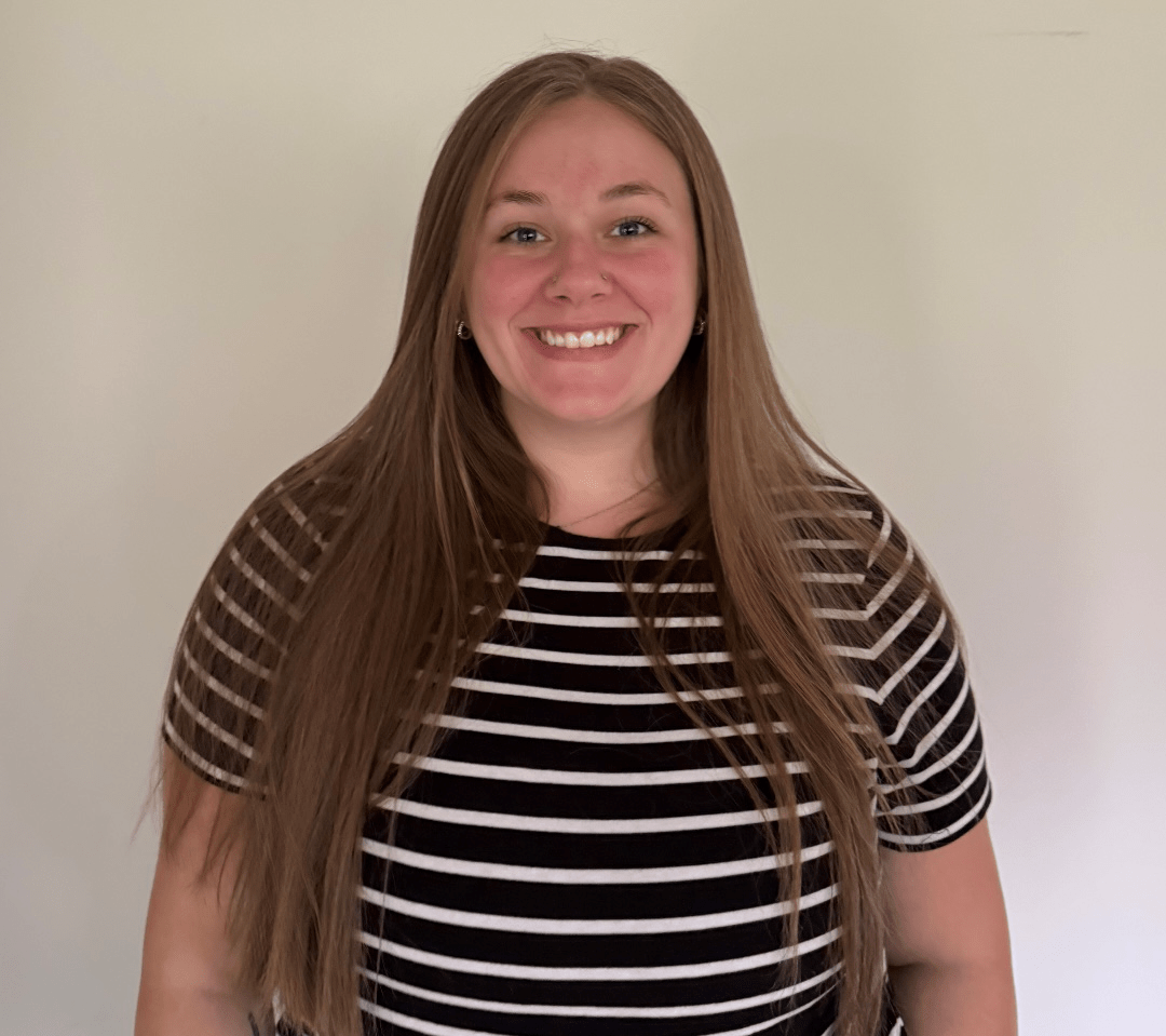 A young woman with long, straight light brown hair smiles at the camera. She is wearing a black and white horizontal striped shirt and standing against a plain light-colored wall.