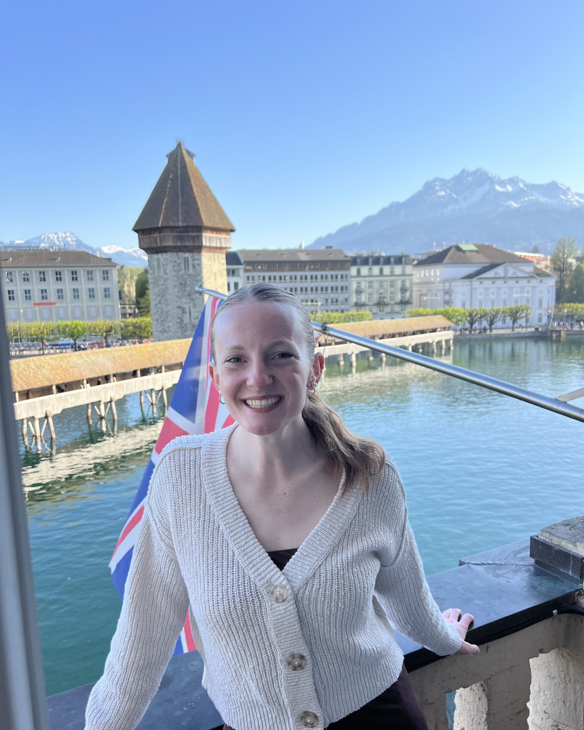 A smiling woman with light brown hair stands on a balcony above a river. Behind her are a historic wooden bridge, old buildings, a British flag, and snow-capped mountains under a clear blue sky.