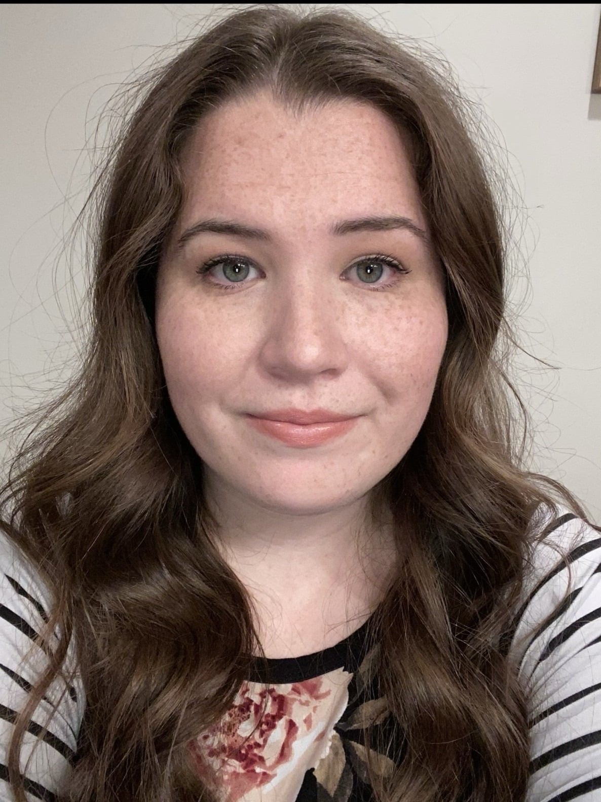 A woman with long, wavy brown hair and light skin is smiling slightly. She is wearing a patterned top with black and white stripes and a floral design. The background is neutral and out of focus.