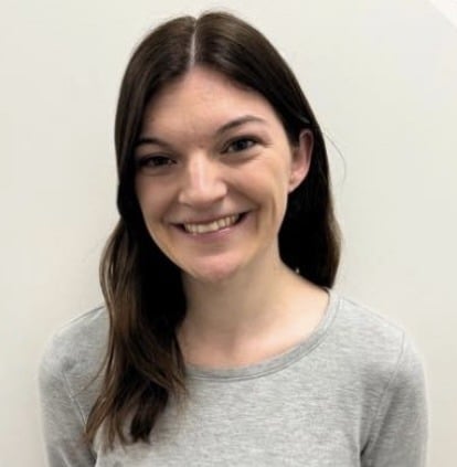 A young woman with straight brown hair, wearing a light gray long-sleeve shirt, smiles at the camera against a plain white background.