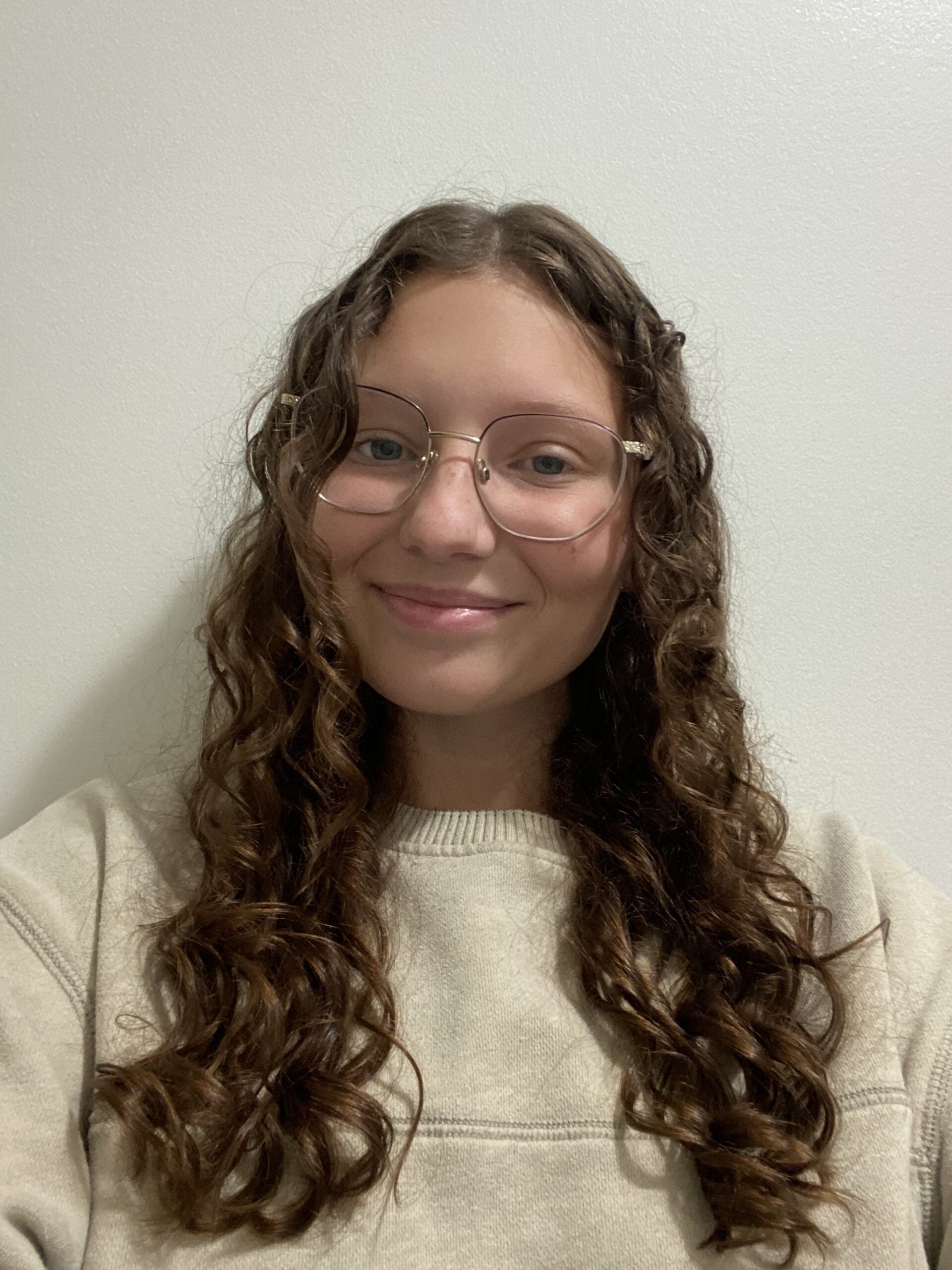 A young person with long, curly brown hair and glasses smiles at the camera. They are wearing a light beige sweater and are posed in front of a plain light-colored wall.