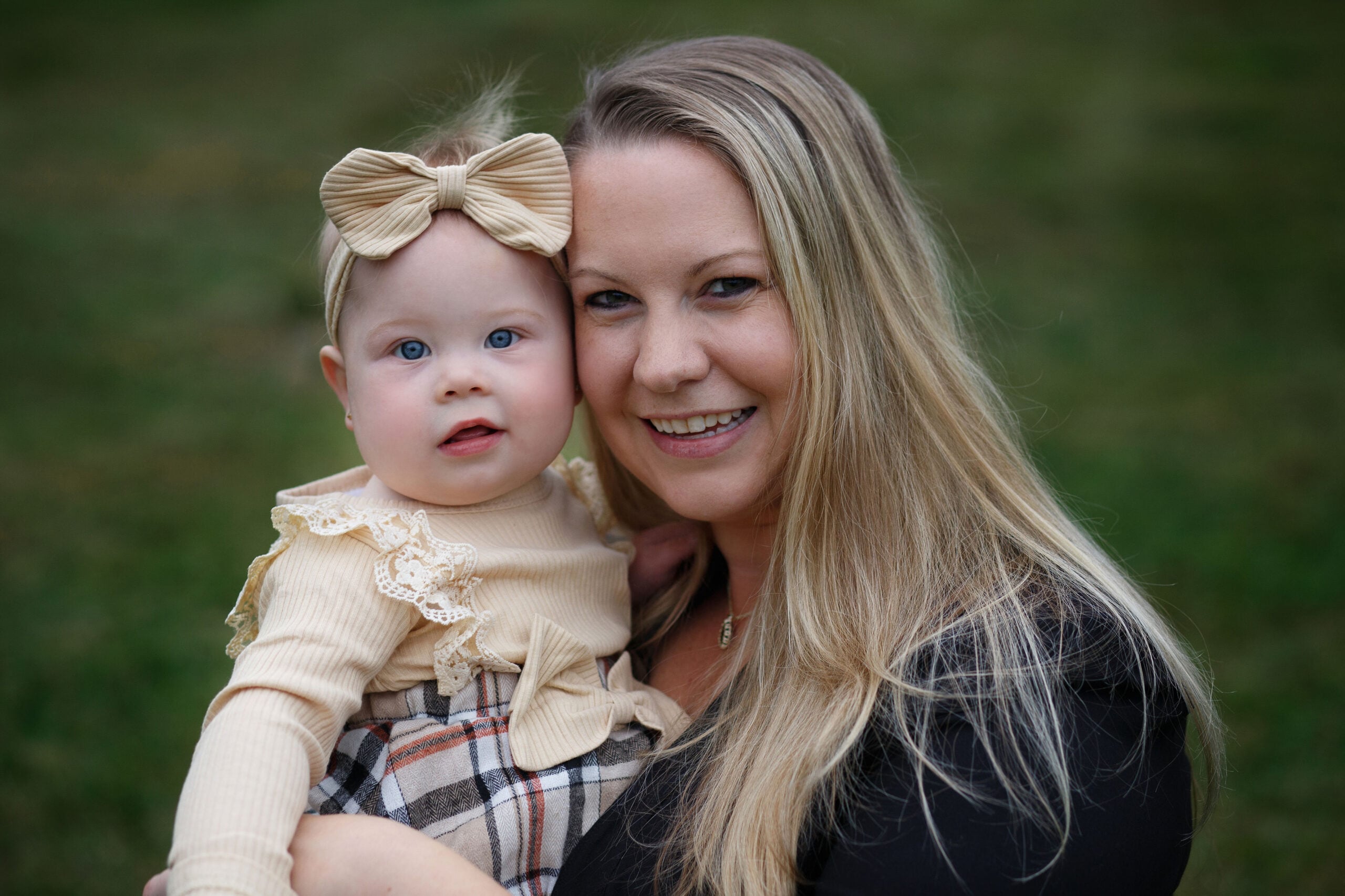 A woman with long blonde hair smiles at the camera while holding a baby girl with a beige bow headband and beige outfit. They are outdoors with a blurred grassy background.