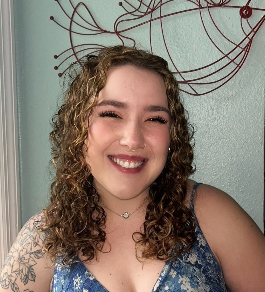 A woman with curly brown hair and a nose ring smiles at the camera. She wears a blue floral dress and a necklace. A decorative red wire art piece hangs on the light green wall behind her.