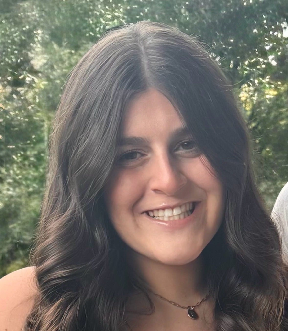 A woman with long, wavy dark hair smiles at the camera in an outdoor setting with green, leafy trees in the background. She is wearing a necklace and a light-colored top.