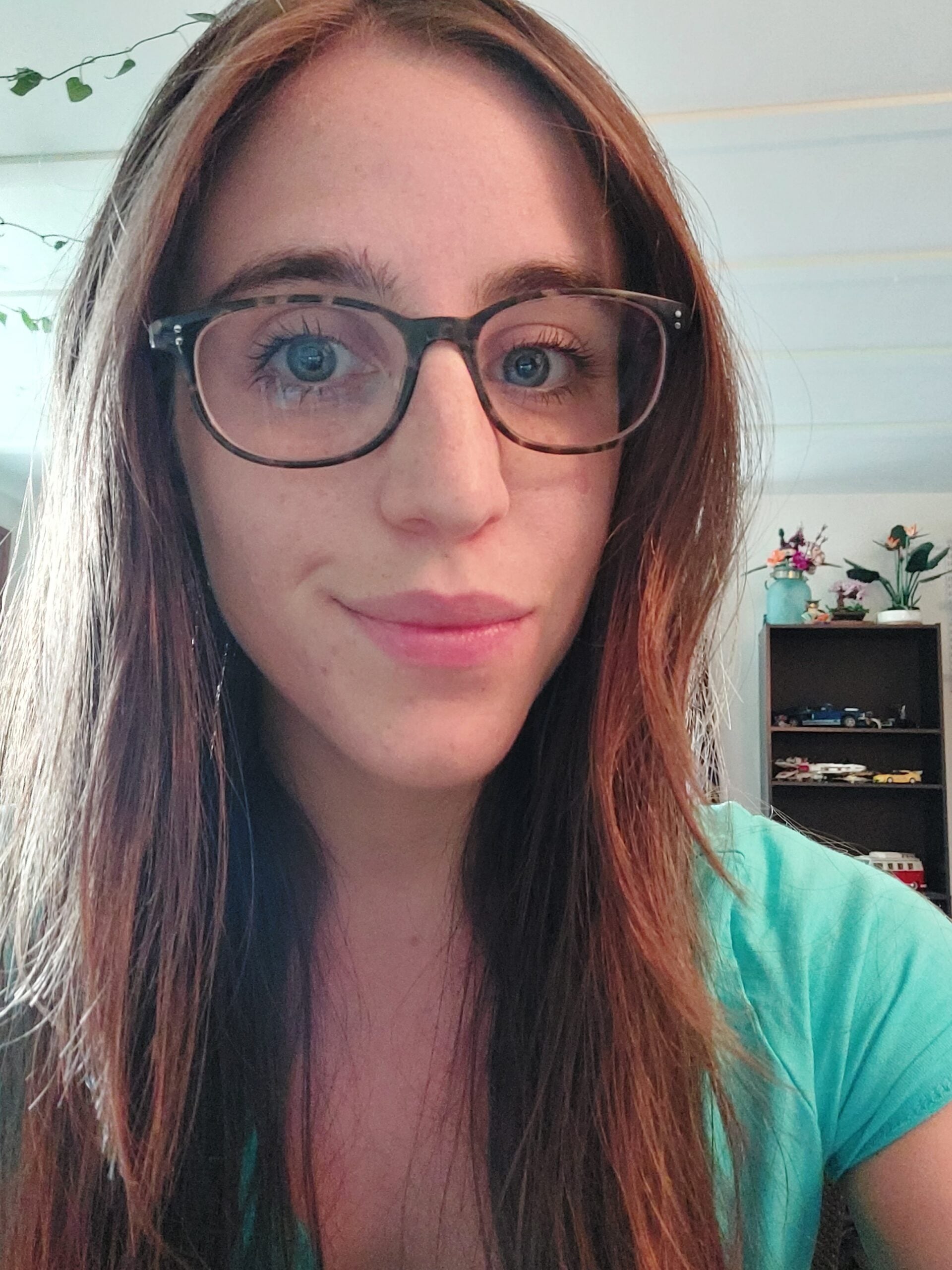 A woman with long brown hair and glasses is smiling slightly at the camera. She is wearing a teal top. In the background, there is a shelf with various objects and plants, and some leafy decorations hang from the ceiling.