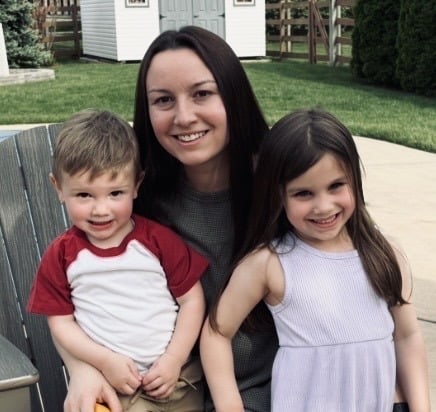 A woman sits outdoors smiling with a young boy in a red and white shirt on her left and a young girl in a light purple dress on her right. They are in a backyard with green grass and a shed in the background.