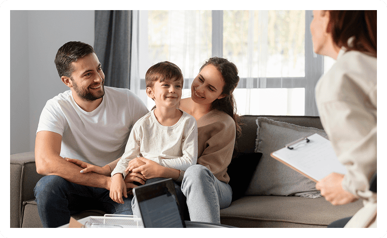 A smiling family—father, mother, and young son—sit together on a couch, engaging with a woman holding a clipboard, possibly during a counseling or consultation session in a bright living room.