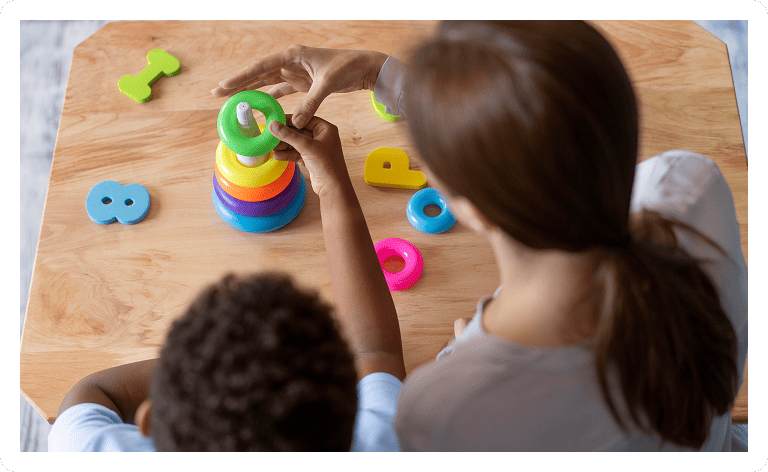 A woman and a child sit at a wooden table, stacking colorful rings onto a peg, with plastic numbers scattered nearby. The activity appears educational and engaging.