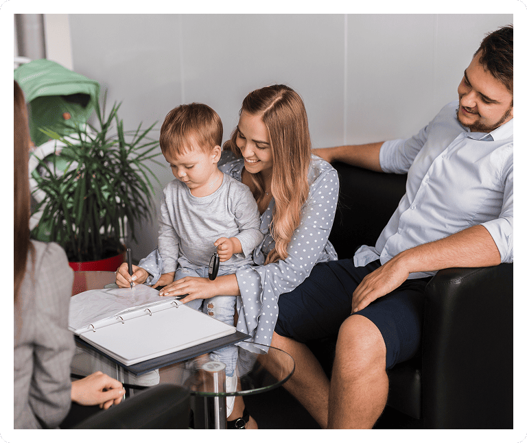A young boy sits on his mother's lap and writes in a binder on a glass table, while his parents smile and watch him. Another person is seated across from them, partially visible.