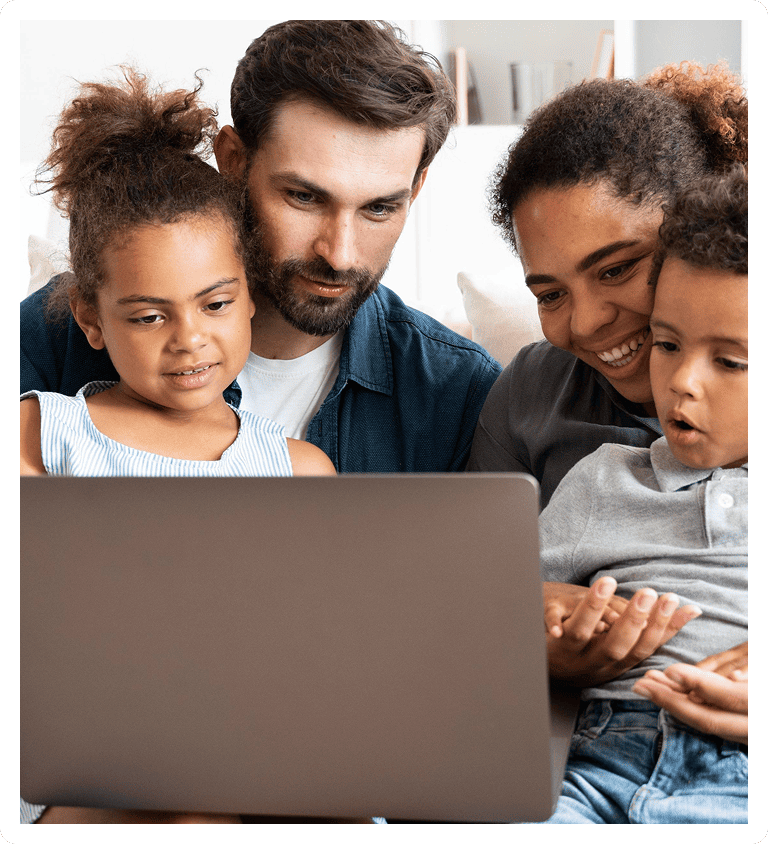 A family of four sits closely together, smiling and looking at a laptop screen. Two adults and two young children appear engaged and happy as they share the experience on the computer.