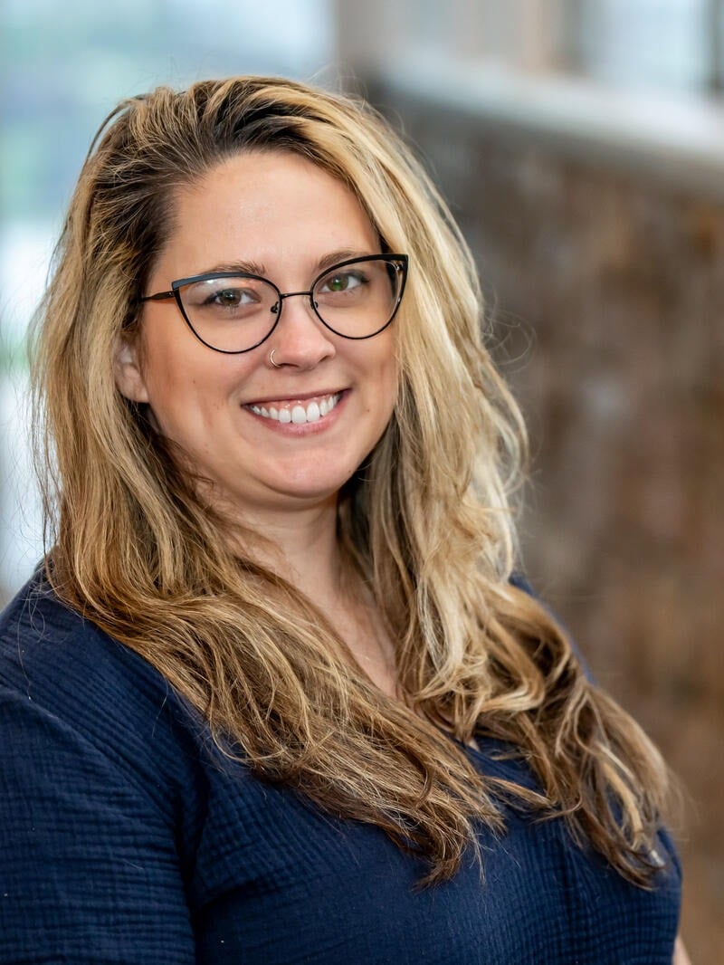 A woman with long, wavy blonde hair and glasses smiles at the camera. She is wearing a dark top and standing indoors with a softly blurred background.