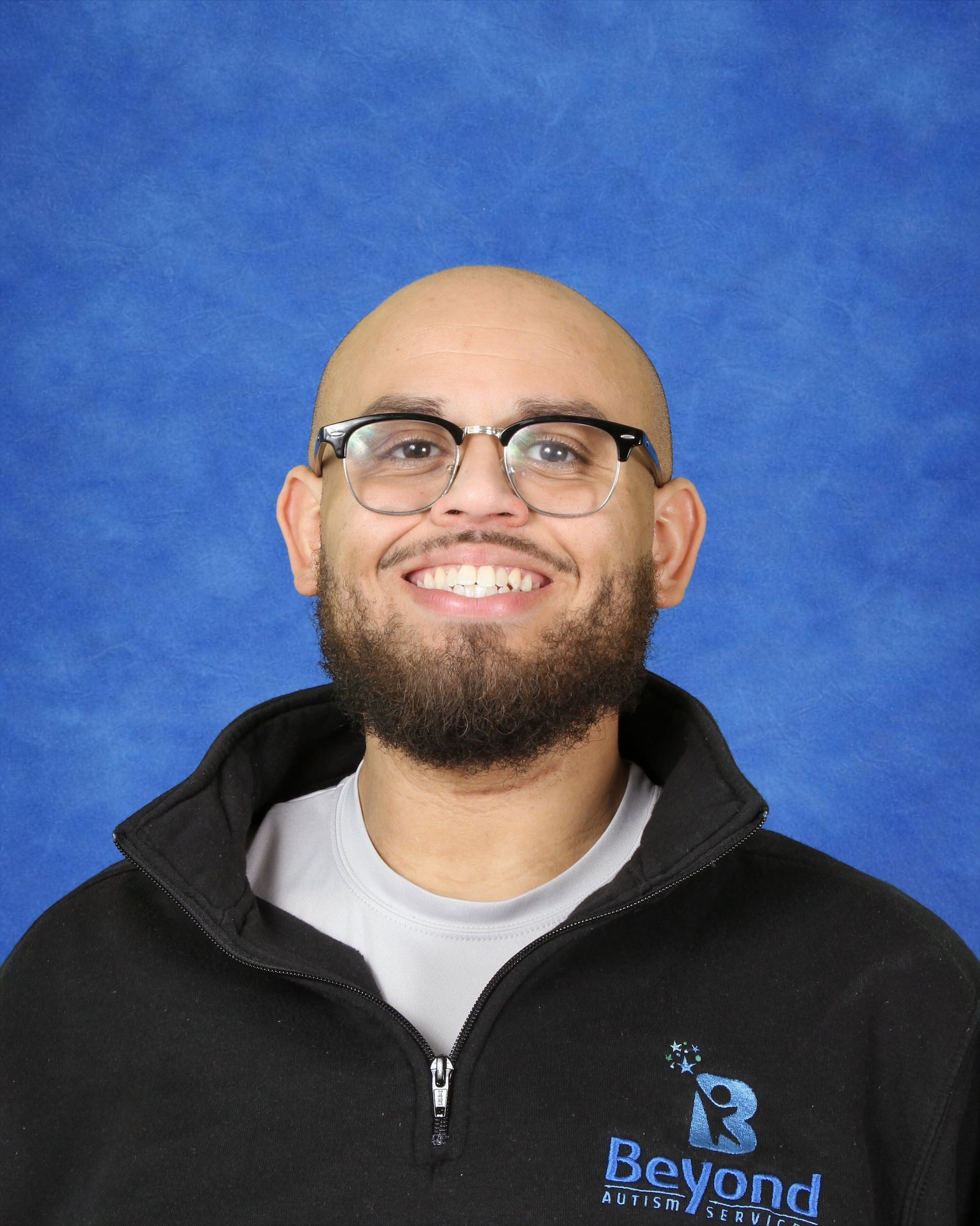 Smiling man with glasses and a beard wearing a black jacket with the "Beyond Autism Services" logo, posed in front of a blue textured background.