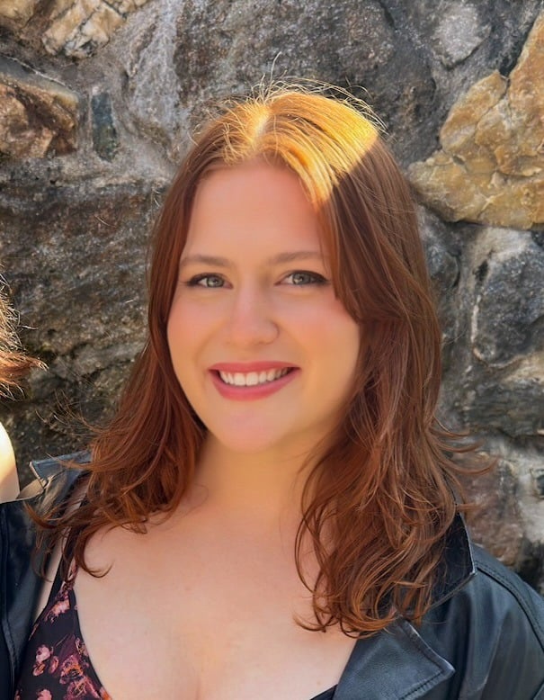 A woman with long, auburn hair and fair skin smiles at the camera. She is wearing a black jacket and a patterned top, standing in front of a stone wall in sunlight.