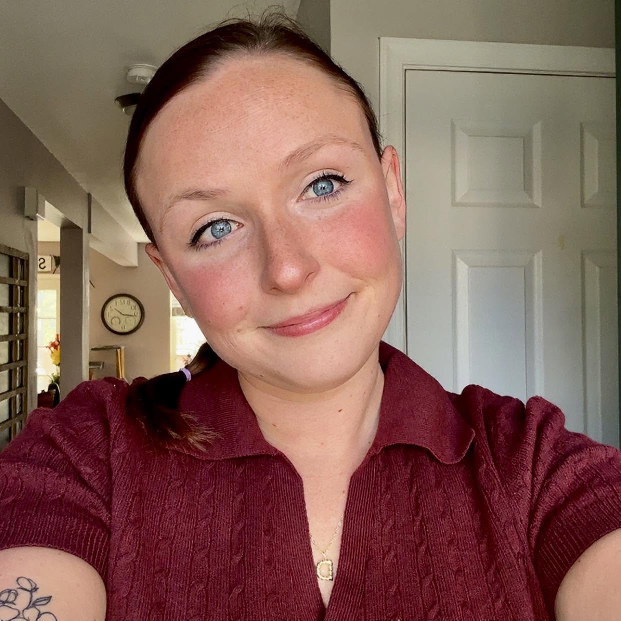 A person with fair skin, blue eyes, and brown hair tied back smiles slightly while wearing a maroon collared shirt. A clock and part of a kitchen are visible in the background.